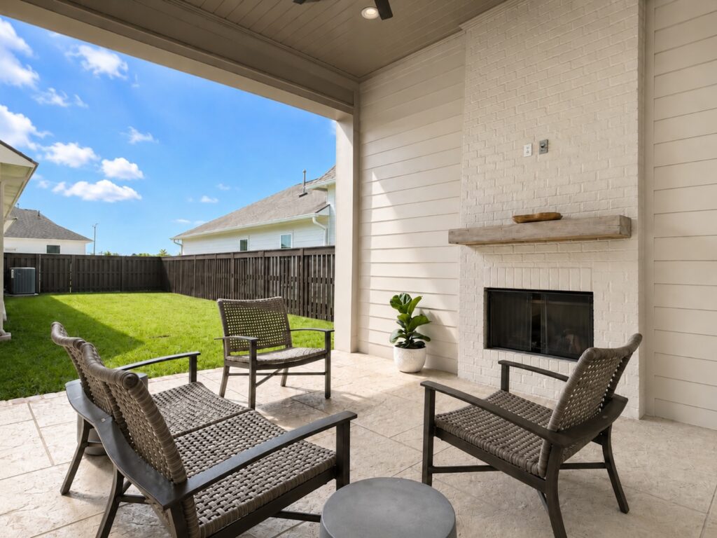 Covered patio with wicker chairs around a small round table, a fireplace on a white brick wall, and a potted plant, overlooking a green backyard under a blue sky.