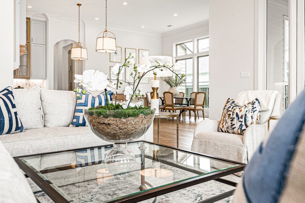 Bright open-concept living room with light gray sofas, blue patterned cushions, a glass coffee table, and a large green plant centerpiece in a glass bowl, with a dining area visible in the background.